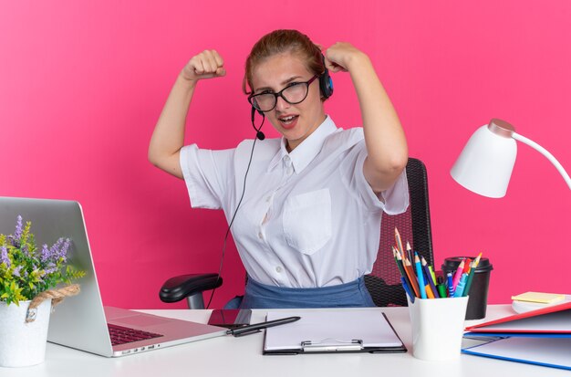 Selbstbewusstes junges blondes Callcenter-Mädchen mit Headset und Brille, das am Schreibtisch mit Arbeitswerkzeugen sitzt und in die Kamera schaut, die starke Geste einzeln auf rosa Wand macht