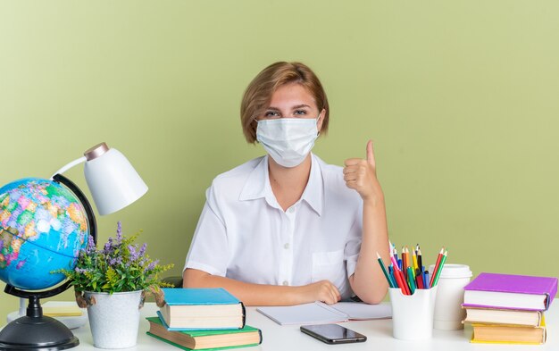 Selbstbewusstes junges blondes Studentenmädchen mit Schutzmaske, das am Schreibtisch mit Schulwerkzeugen sitzt und in die Kamera schaut, die den Daumen isoliert auf der olivgrünen Wand zeigt