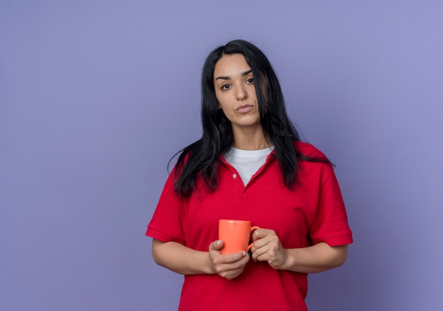 Selbstbewusstes junges brünettes kaukasisches Mädchen, das rotes Hemd trägt, hält Tasse lokalisiert auf lila Wand