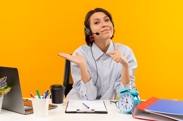 Selbstbewusstes junges Callcenter-Mädchen, das ein Headset trägt, das am Schreibtisch sitzt und leere Hand zeigt und auf es lokalisiert auf Orange zeigt