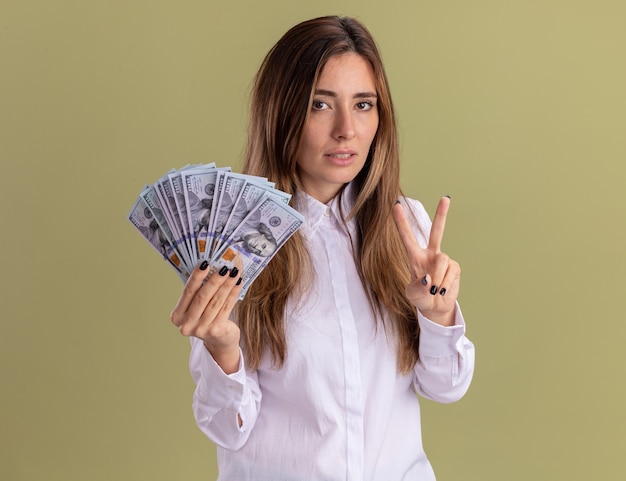 Selbstbewusstes junges hübsches kaukasisches Mädchen hält Geld und gestikuliert Siegeshandzeichen