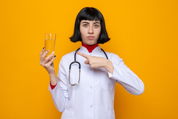 Selbstbewusstes junges hübsches kaukasisches Mädchen in Arztuniform mit Stethoskop, das ein Glas Wasser hält