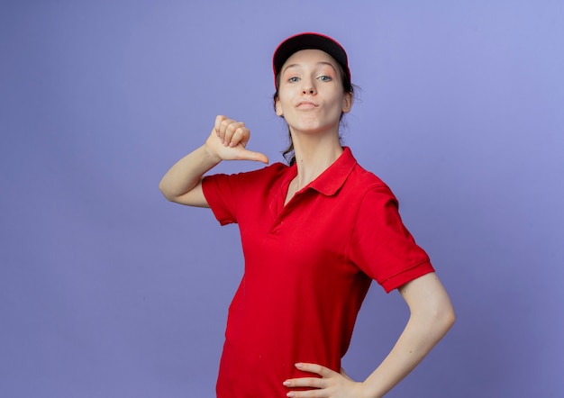 Selbstbewusstes junges hübsches Liefermädchen, das rote Uniform und Kappe trägt, die Hand auf Taille setzen und auf sich selbst lokalisiert auf lila Hintergrund mit Kopienraum zeigt