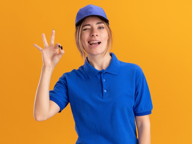 Selbstbewusstes junges hübsches Liefermädchen in Uniform blinkt Auge und gestikuliert ok Handzeichen auf Orange