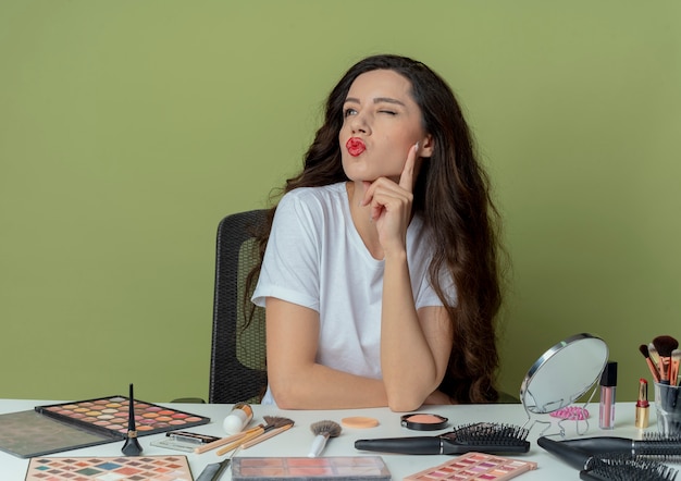 Selbstbewusstes junges hübsches mädchen, das am make-up-tisch mit make-up-tools sitzt und die hand unter das kinn legt und auf die seite zwinkert und eine kussgeste auf olivgrünem grünraum macht?