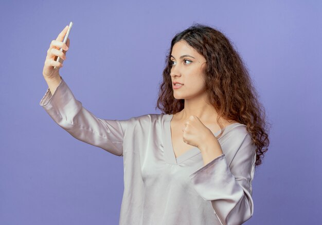 Selbstbewusstes junges hübsches Mädchen nehmen ein Selfie und halten Faust lokalisiert auf blauer Wand