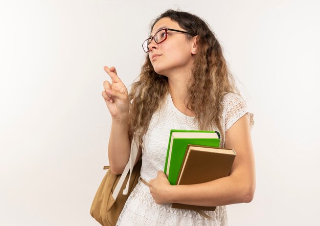 Selbstbewusstes junges hübsches Schulmädchen, das Brille und Rückentasche hält Bücher hält, die Seitenkreuzungsfinger lokalisiert auf Weiß mit Kopienraum betrachten