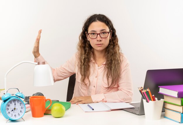 Selbstbewusstes junges hübsches Schulmädchen, das eine Brille trägt, die am Schreibtisch mit Schulwerkzeugen sitzt und ihre Hausaufgaben macht, gestikuliert an der Seite lokalisiert auf Weiß