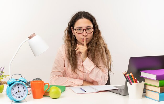 Selbstbewusstes junges hübsches Schulmädchen, das eine Brille trägt, die mit Schulwerkzeugen am Schreibtisch sitzt und ihre Hausaufgaben macht, die Stille gestikuliert lokalisiert auf Weiß