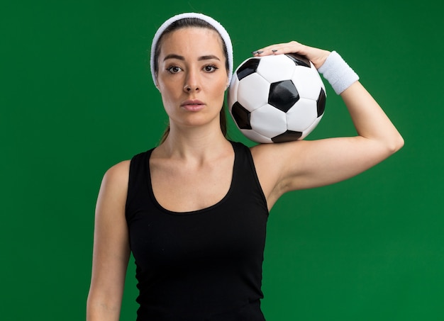 Selbstbewusstes junges hübsches sportliches Mädchen mit Stirnband und Armbändern, das Fußball auf der Schulter isoliert auf grüner Wand hält