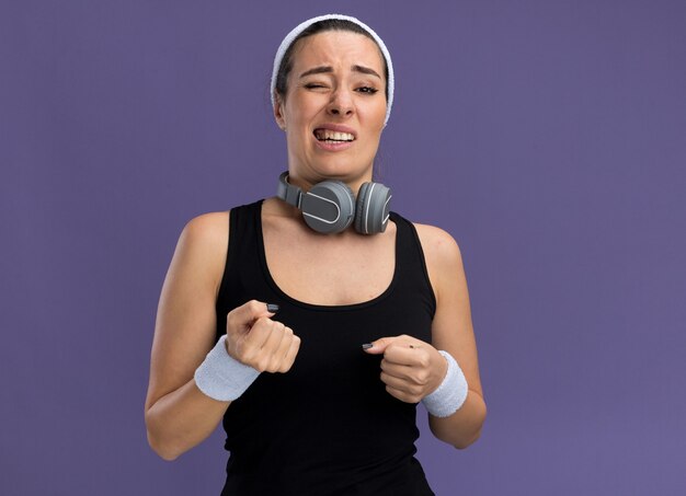 Selbstbewusstes junges hübsches sportliches Mädchen mit Stirnband und Armbändern mit Kopfhörern um den Hals, die Fäuste mit einem geschlossenen Auge geschlossen auf lila Wand mit Kopienraum