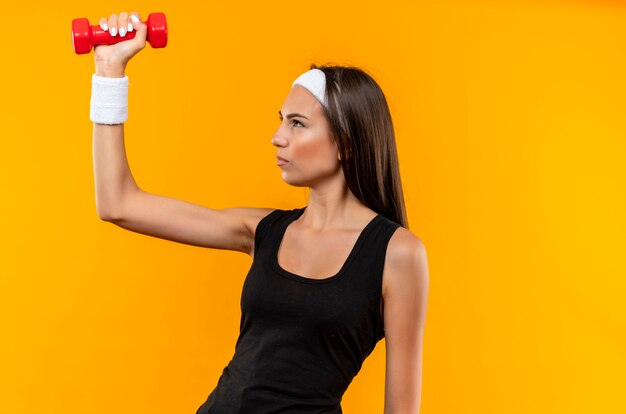 Selbstbewusstes junges hübsches sportliches Mädchen mit Stirnband und Armband, das Hantel einzeln auf oranger Wand mit Kopierraum anhebt und betrachtet