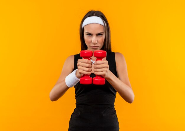 Selbstbewusstes junges hübsches sportliches Mädchen mit Stirnband und Armband, das Hanteln ausstreckt und isoliert auf oranger Wand aussieht