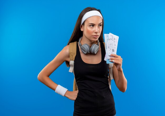 Selbstbewusstes junges hübsches sportliches Mädchen mit Stirnband und Armband und Rückentasche und Kopfhörern am Hals mit Flugtickets auf der Seite isoliert auf blauer Wand
