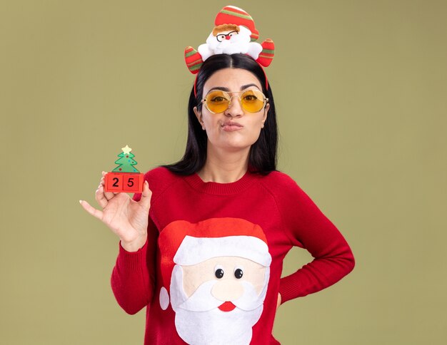 Selbstbewusstes junges kaukasisches Mädchen, das Weihnachtsmann-Stirnband und Pullover mit Brille trägt, die Weihnachtsbaumspielzeug mit Datum hält, das Hand auf Taille hält und Kamera lokalisiert auf olivgrünem Hintergrund betrachtet
