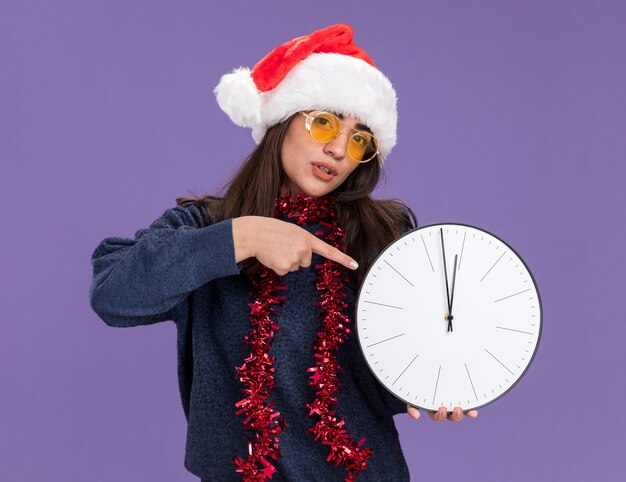 selbstbewusstes junges kaukasisches Mädchen in Sonnenbrille mit Weihnachtsmütze und Girlande um Hals hält und zeigt auf Uhr lokalisiert auf lila Hintergrund mit Kopienraum