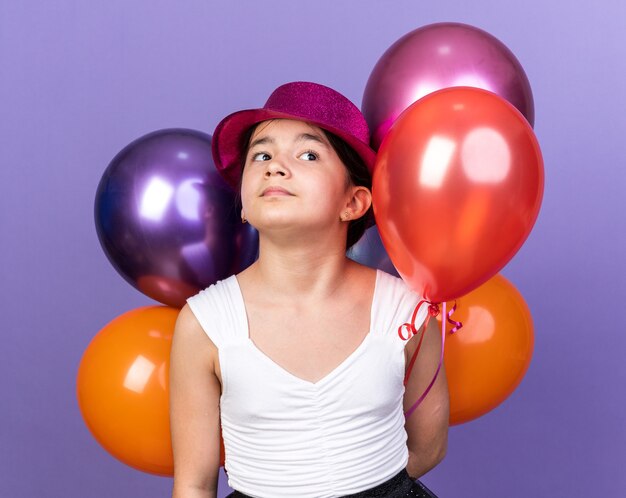 selbstbewusstes junges kaukasisches Mädchen mit violettem Partyhut, der Heliumballons hält und die Seite isoliert auf lila Wand mit Kopienraum betrachtet