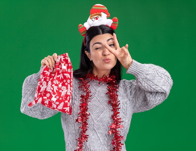 selbstbewusstes junges kaukasisches Mädchen mit Weihnachtsmann-Stirnband und Lametta-Girlande um den Hals, das einen Weihnachtsgeschenksack hält und ein Friedenszeichen vor dem Auge macht, das isoliert auf grüner Wand zwinkert?