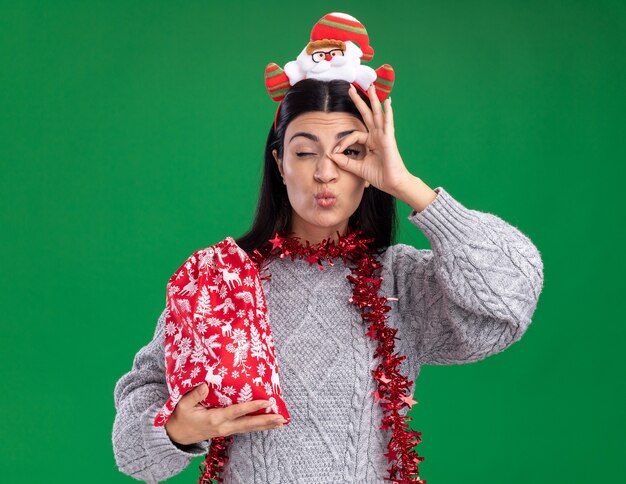 selbstbewusstes junges kaukasisches Mädchen mit Weihnachtsmann-Stirnband und Lametta-Girlande um den Hals, das einen Weihnachtsgeschenksack hält