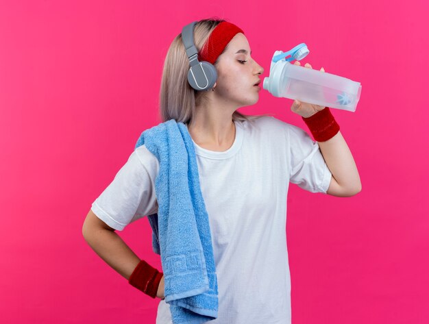 Selbstbewusstes junges kaukasisches sportliches Mädchen mit Hosenträgern an Kopfhörern, das Stirnband und Armbänder trägt, hält ein Handtuch auf der Schulter und trinkt aus einer Wasserflasche