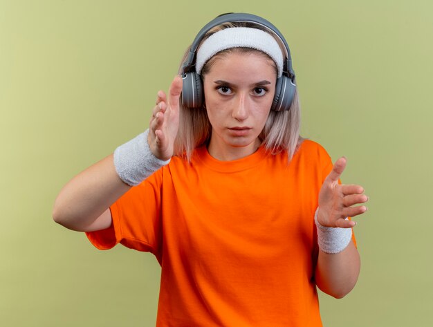 Selbstbewusstes junges kaukasisches sportliches Mädchen mit Hosenträgern auf Kopfhörern mit Stirnband und Armbändern hält die Hände direkt an die Kamera