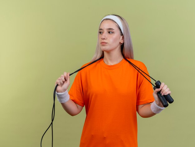 Selbstbewusstes junges, kaukasisches, sportliches Mädchen mit Hosenträgern, das Stirnband und Armbänder trägt, hält Springseil um den Hals und schaut auf die Seite