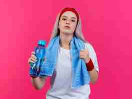 Kostenloses Foto selbstbewusstes junges kaukasisches sportliches mädchen mit hosenträgern und mit handtuch um den hals, das stirnband und armbänder mit wasserflasche trägt