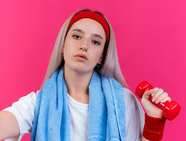 Selbstbewusstes junges kaukasisches sportliches Mädchen mit Hosenträgern und mit Handtuch um den Hals, das Stirnband und Armbänder trägt, hält Hantel mit Blick auf die Kamera