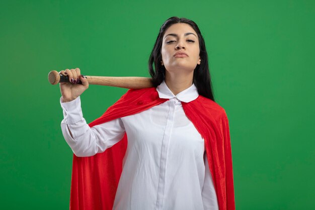 Selbstbewusstes junges kaukasisches Superheldenmädchen, das Baseballschläger auf ihrer Schulter hält und Kamera lokalisiert auf grünem Hintergrund betrachtet