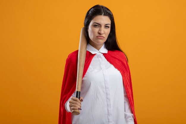 Selbstbewusstes junges kaukasisches Superheldenmädchen, das Baseballschläger hält, der Kamera lokalisiert auf orange Hintergrund mit Kopienraum betrachtet
