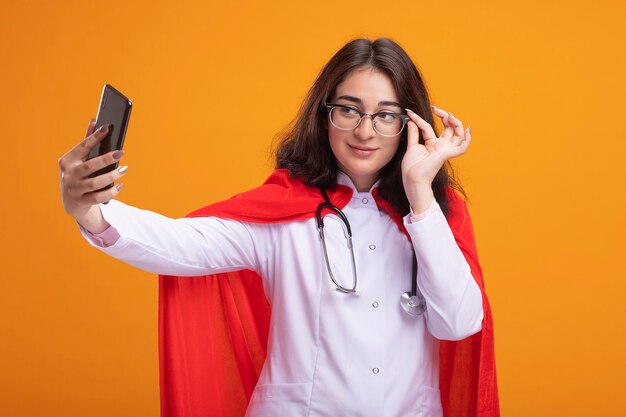 Selbstbewusstes junges kaukasisches Superheldenmädchen in rotem Umhang mit Arztuniform und Stethoskop mit Brille, die eine Brille greift und ein Selfie macht