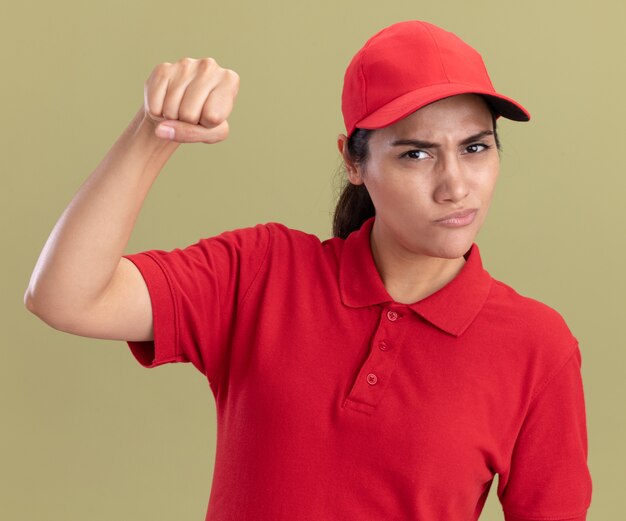 Selbstbewusstes junges Liefermädchen, das Uniform mit Kappe trägt, macht starke Geste lokalisiert auf olivgrüner Wand