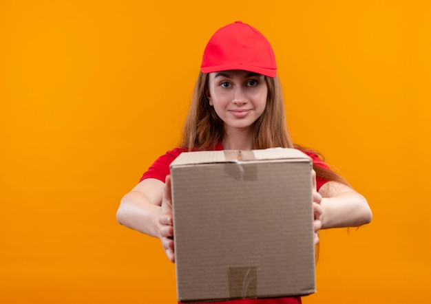 Selbstbewusstes junges Liefermädchen in der roten Uniform, die Box auf lokalisiertem orange Raum mit Kopienraum streckt