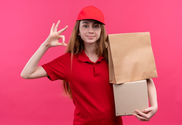 Selbstbewusstes junges Liefermädchen in der roten Uniform, die Kisten hält und ok Zeichen auf lokalisiertem rosa Raum tut