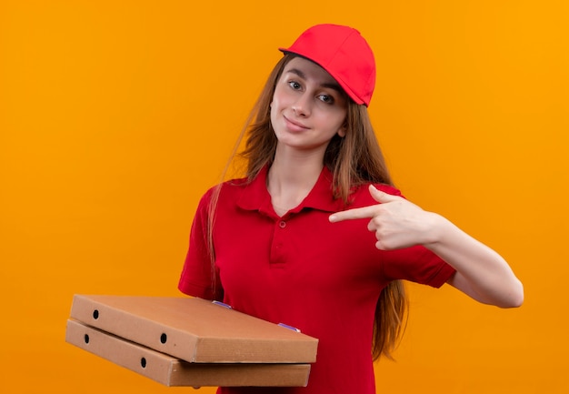 Selbstbewusstes junges Liefermädchen in der roten Uniform, die Pakete hält und auf sie auf lokalisiertem orangefarbenem Raum zeigt