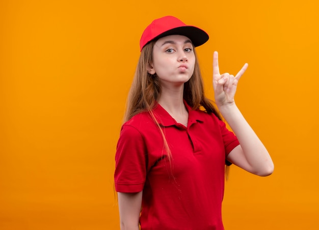 Selbstbewusstes junges Liefermädchen in der roten Uniform, die Rockzeichen auf lokalisiertem orangefarbenem Raum mit Kopienraum tut