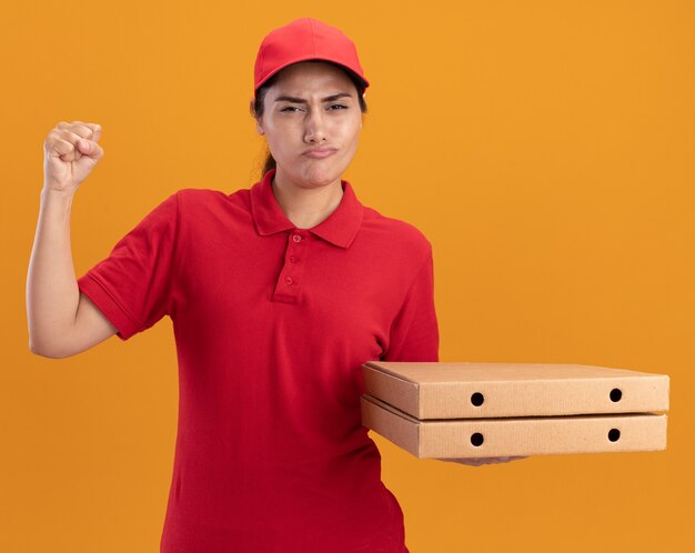 Selbstbewusstes junges Liefermädchen in Uniform und Mütze, das Pizzakartons hält und die Faust einzeln auf oranger Wand hebt