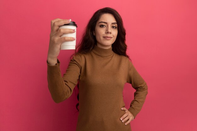 Selbstbewusstes junges schönes Mädchen, das Rollkragenpullover trägt, der Tasse Kaffee lokalisiert auf rosa Wand hält