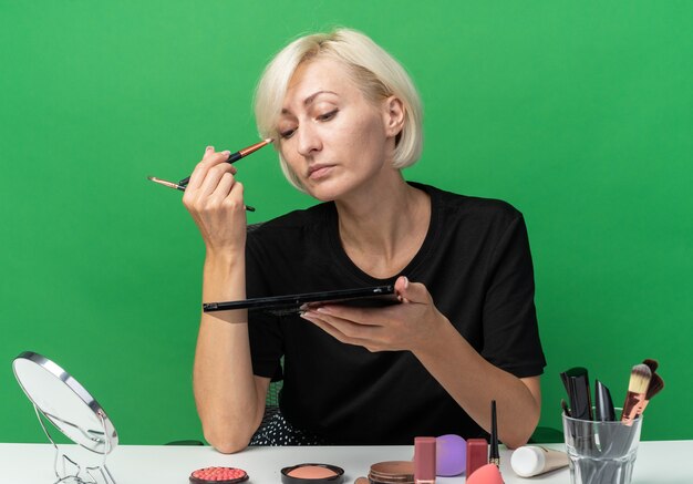 Selbstbewusstes junges schönes Mädchen sitzt am Tisch mit Make-up-Tools, die Lidschatten mit Make-up-Pinsel auf grüner Wand auftragen