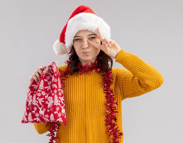 Selbstbewusstes junges slawisches Mädchen mit Weihnachtsmütze und mit Girlande um Hals, die Weihnachtsgeschenktüte und gestikulierendes Siegeszeichen lokalisiert auf weißem Hintergrund mit Kopienraum hält