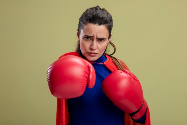 Selbstbewusstes junges Superheldenmädchen, das Boxhandschuhe trägt, die Hand an der Kamera lokalisiert auf olivgrünem Hintergrund halten