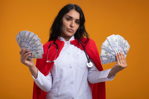 Selbstbewusstes junges Superheldenmädchen, das medizinisches Gewand mit Stethoskop hält, das Geld lokalisiert auf orange Wand hält