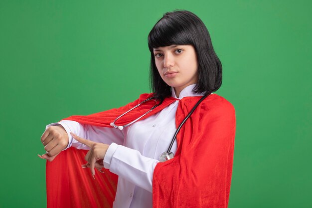 Selbstbewusstes junges Superheldenmädchen, das Stethoskop mit medizinischem Gewand und Umhang trägt, der Armbanduhrgeste zeigt, lokalisiert auf grüner Wand