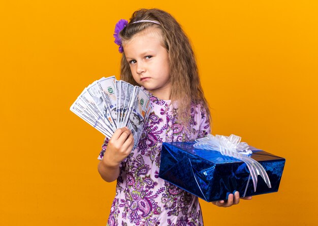 selbstbewusstes kleines blondes Mädchen, das Geschenkbox und Geld lokalisiert auf orange Wand mit Kopienraum hält