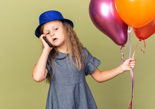 Selbstbewusstes kleines blondes Mädchen mit blauem Partyhut, der Heliumballons hält und telefoniert, isoliert auf olivgrüner Wand mit Kopierraum