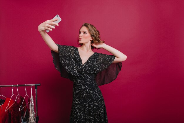 Selbstbewusstes Mädchen mit küssendem Gesichtsausdruck, der Selfie in ihrer Umkleidekabine macht. Stilvolle Frau, die sich in der Nähe von Kleiderbügeln fotografiert.