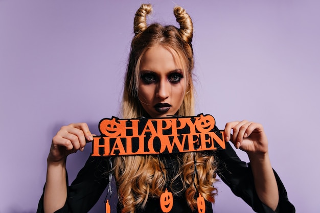 Selbstbewusstes Mädchen mit unheimlichem schwarzem Make-up, das vor Partei aufwirft. ernsthafte blonde Frau im Vampirkostüm, das Halloween feiert.