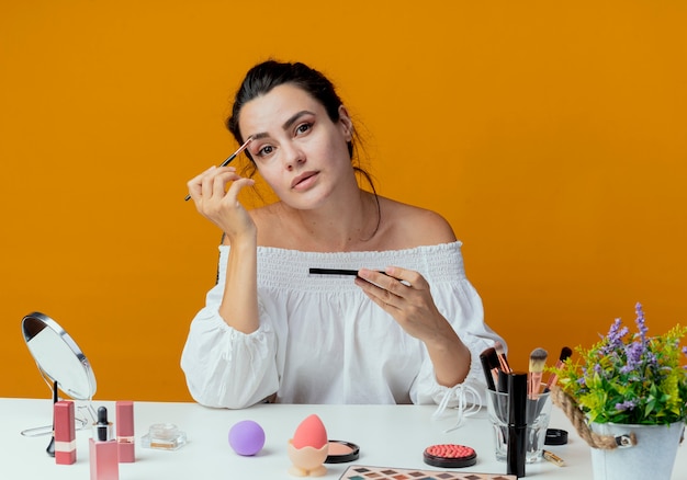 Selbstbewusstes schönes Mädchen sitzt am Tisch mit Make-up-Werkzeugen, die Make-up-Pinsel und Lidschatten-Palette halten, die lokal auf orange Wand schauen