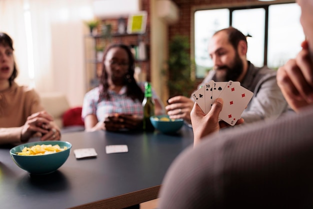 Selektiver Fokus eines Mannes, der Karten in der Hand hält, während er zu Hause mit Freunden spielt. Nachdenkliche Person, die über den nächsten Schritt nachdenkt, während sie Gesellschaftsspiele spielt und Snacks und Getränke genießt.