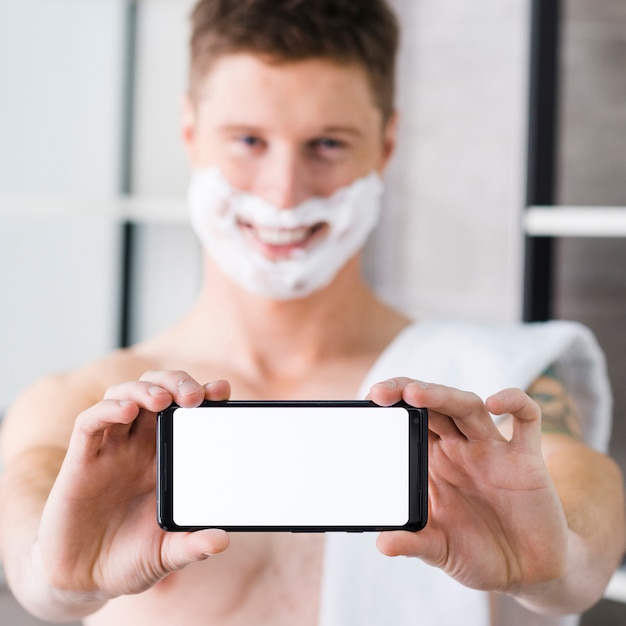 Selektiver Fokus eines Mannes mit dem Rasierschaum auf seinem Gesicht, das leeres weißes intelligentes Telefon in Richtung zur Kamera zeigt